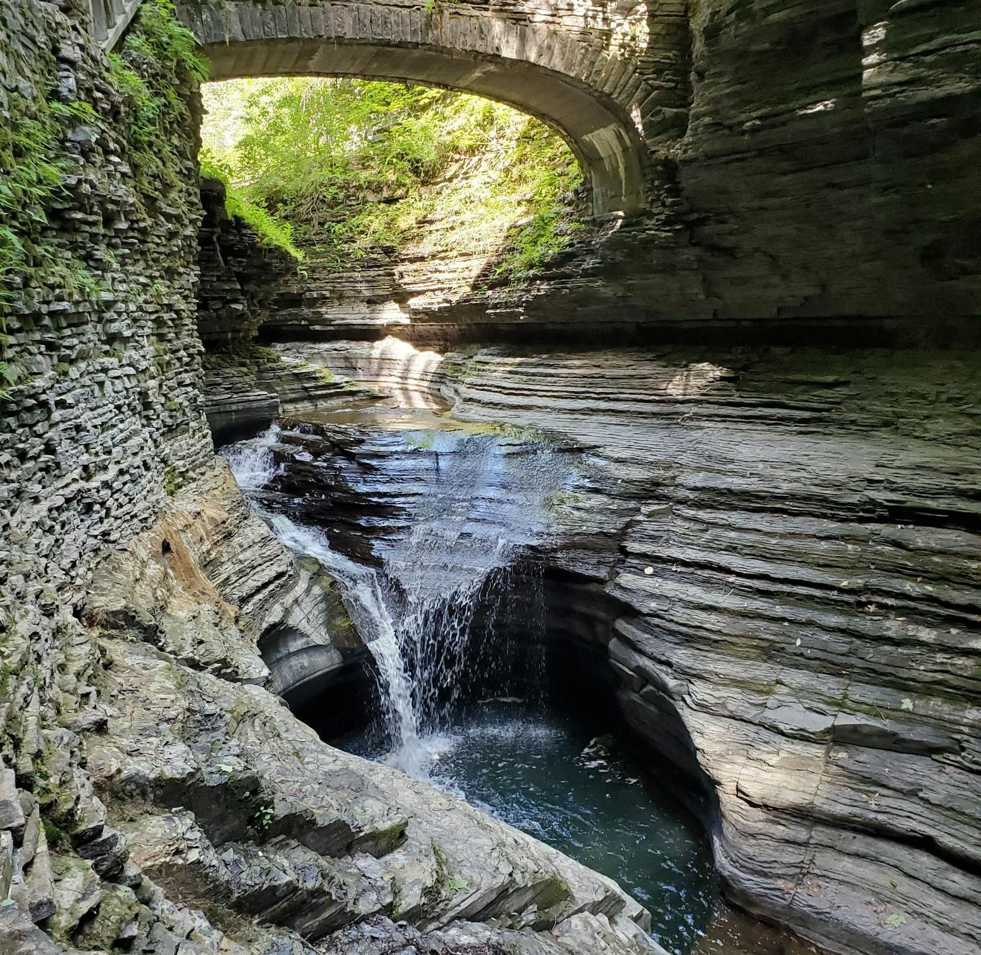 There is a waterfall in the middle of a canyon under a bridge.