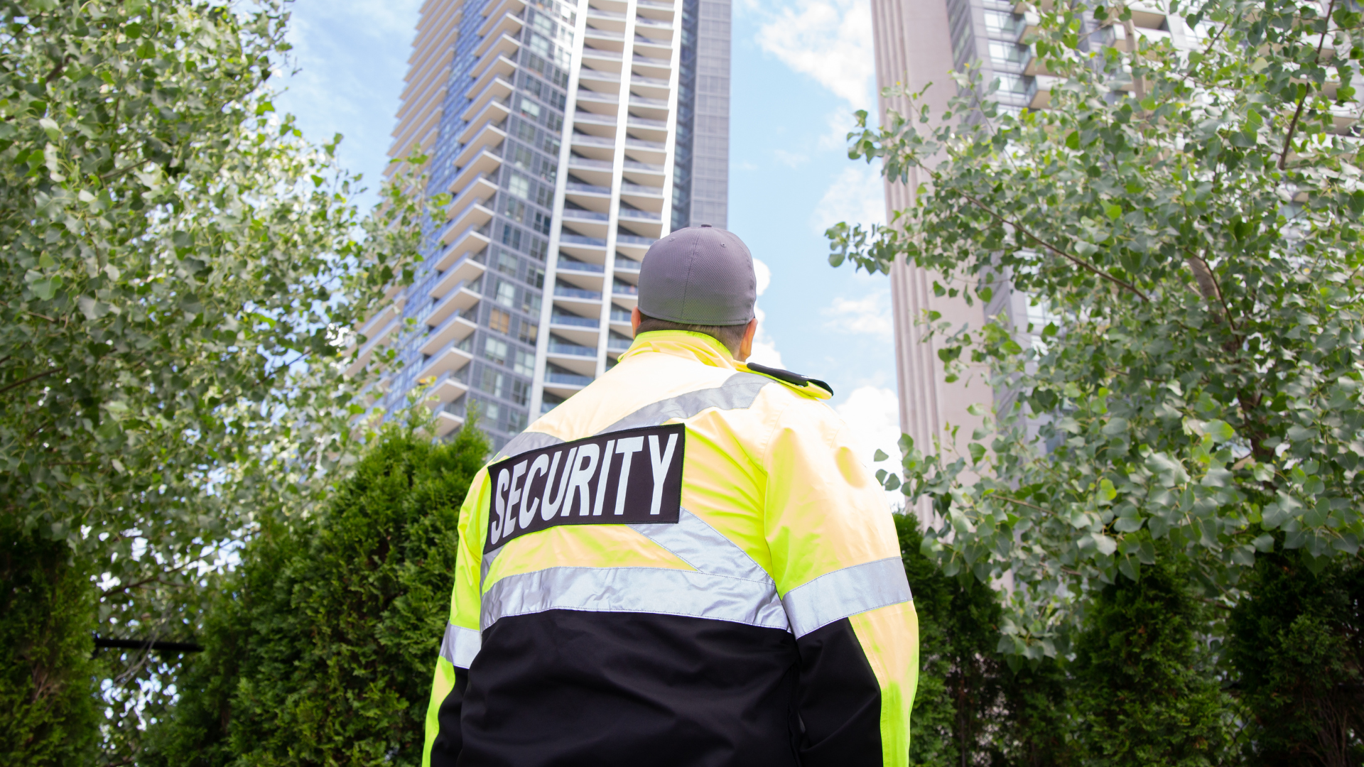 Security guard, yellow and black jacket, looks up at a tall building between trees, outdoors.