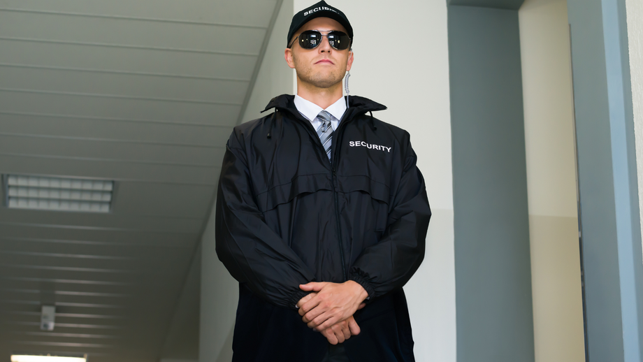 Security guard in black uniform, sunglasses, and cap, standing indoors with hands clasped.
