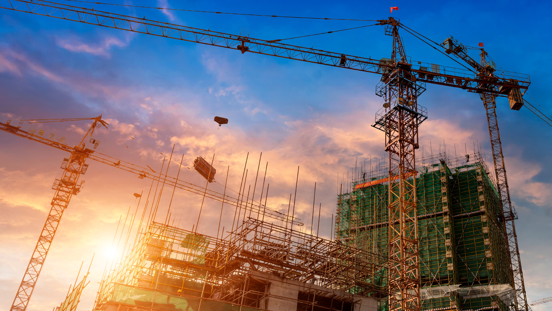 Construction site with cranes against a sunset, building rising, orange and blue sky.