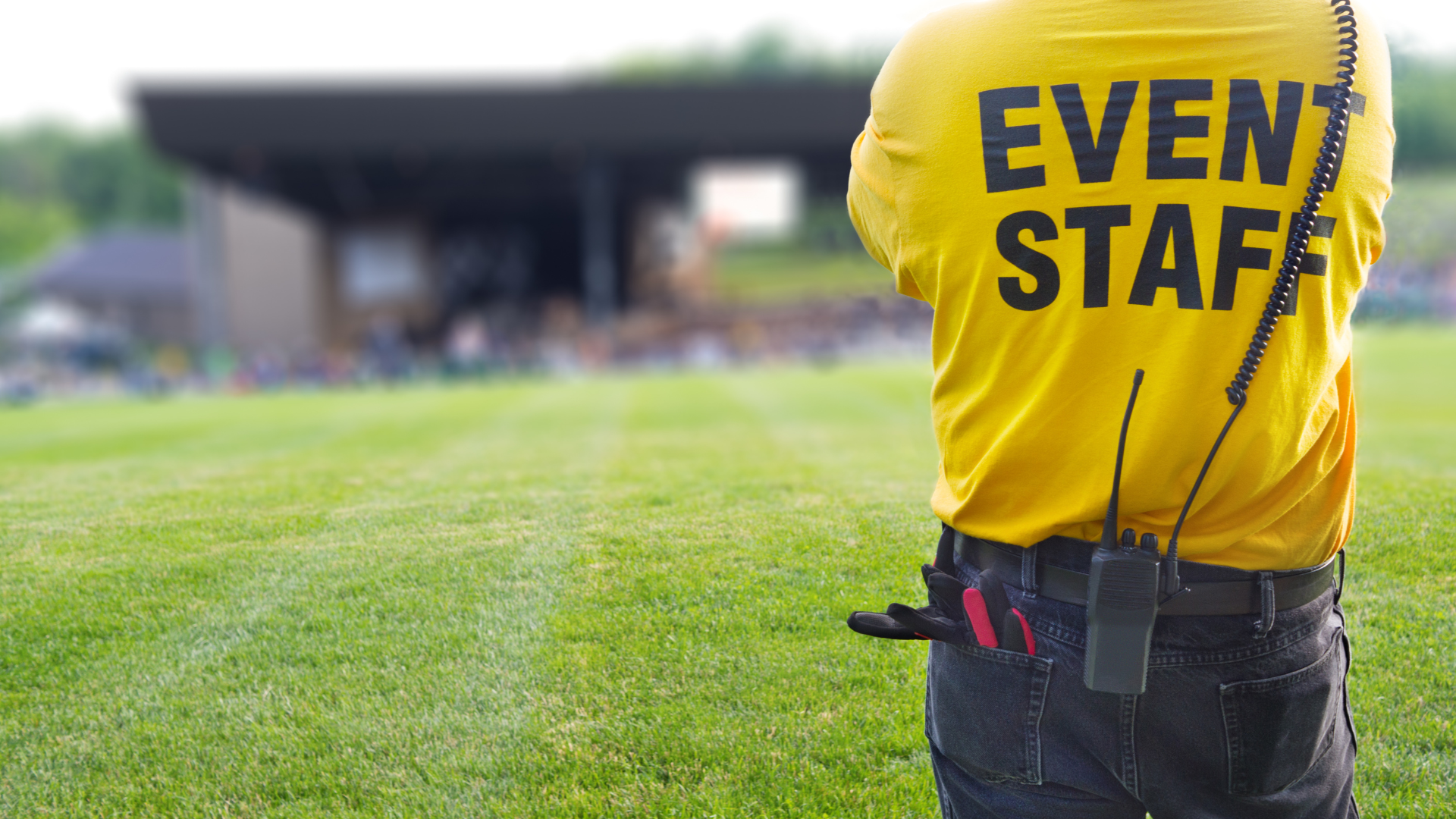 Event staff member in yellow shirt with back to camera; field and stage in background.