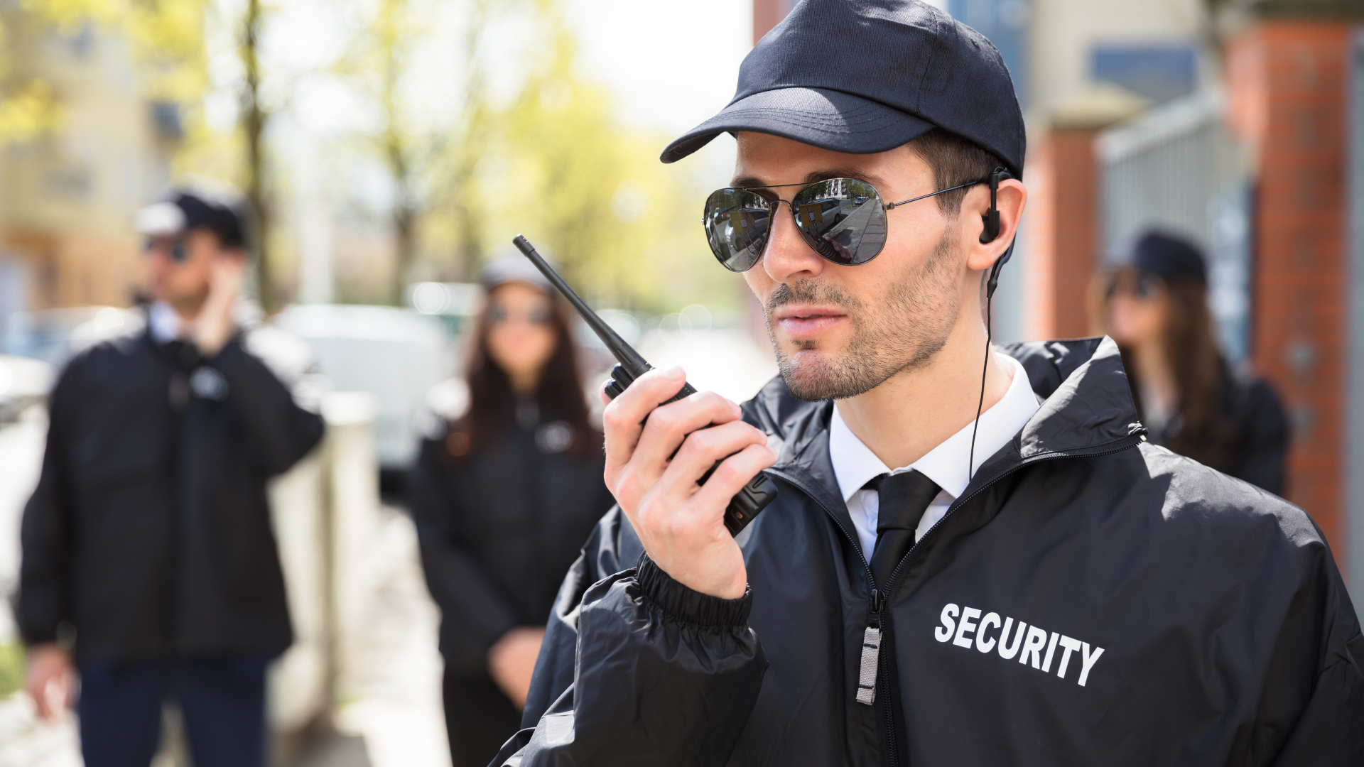 Security guard in black uniform with radio, sunglasses, cap, and earpiece; other guards in background.