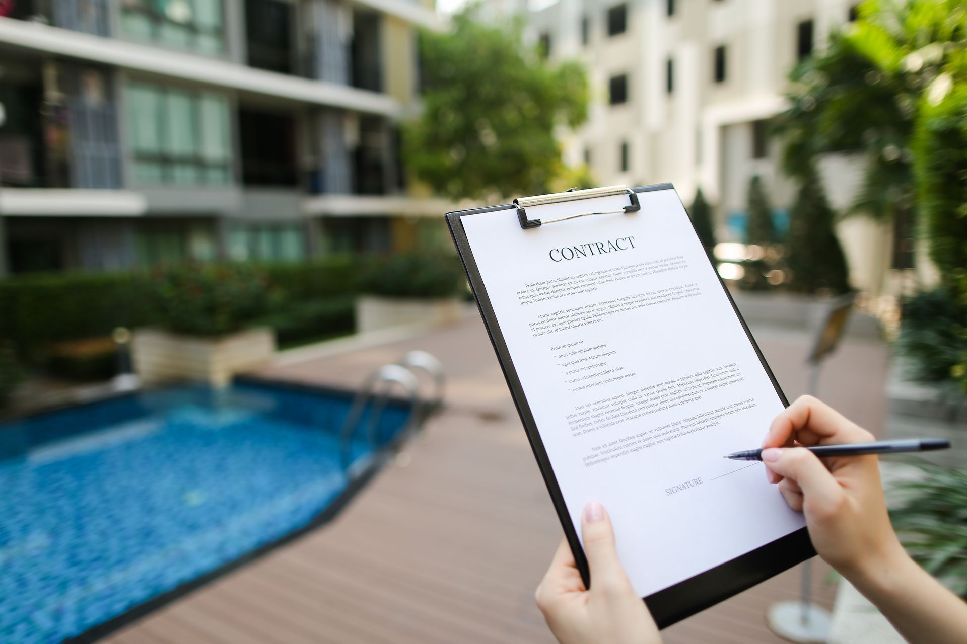 Person signing a contract outdoors by a pool; condos in the background.