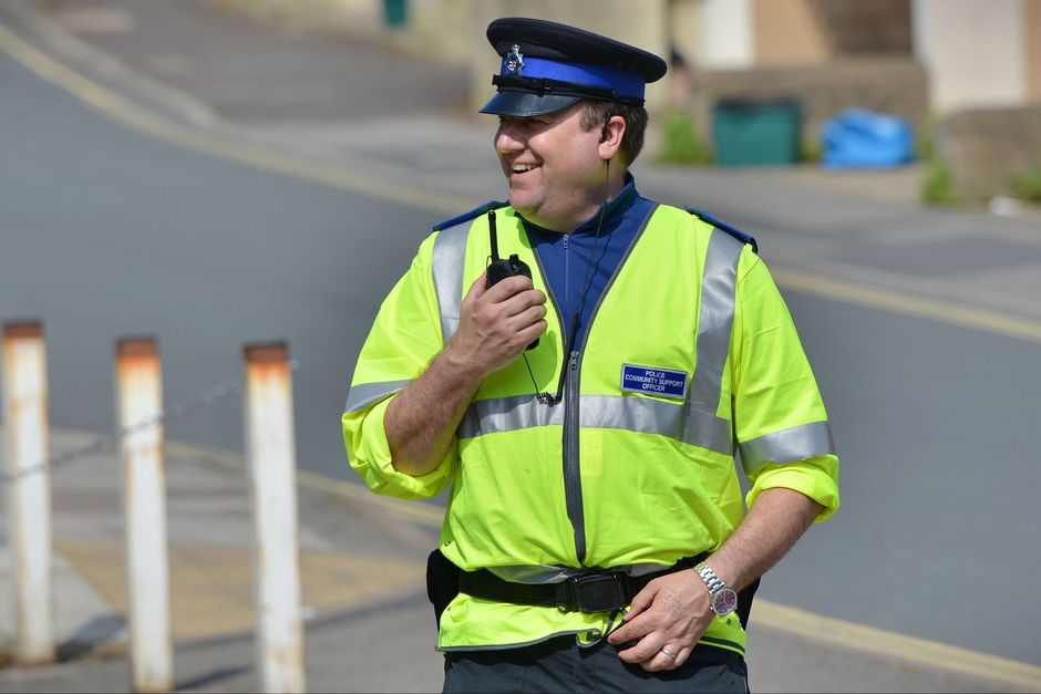 Police officer in yellow vest holding a radio, smiling, on a street.