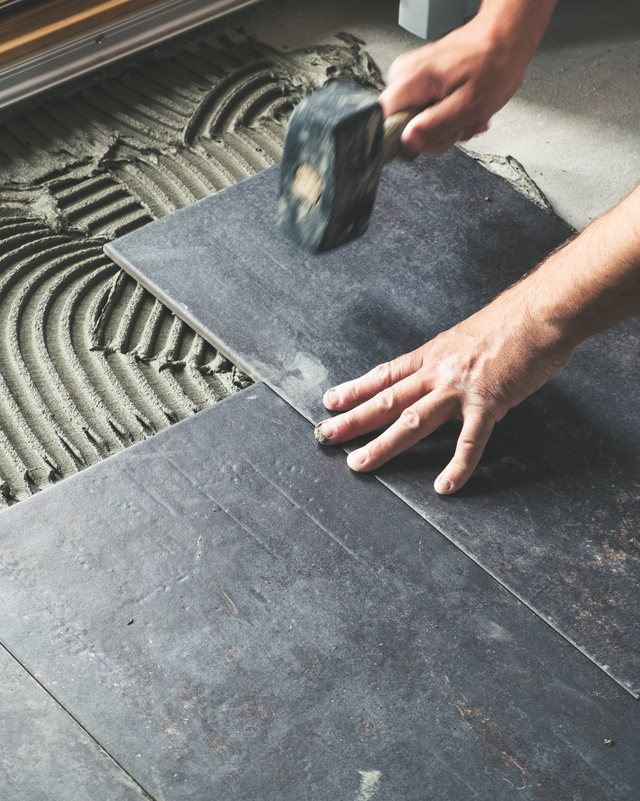 Hands laying dark floor tiles with a mallet over adhesive on the subfloor