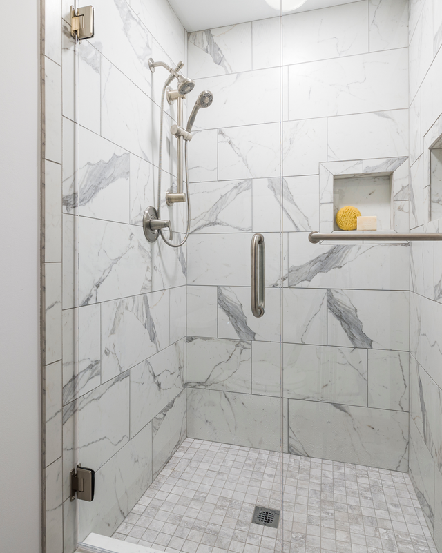 Glass shower with marble tile walls, handheld showerhead, grab bar, and built-in shelf with a yellow bottle.