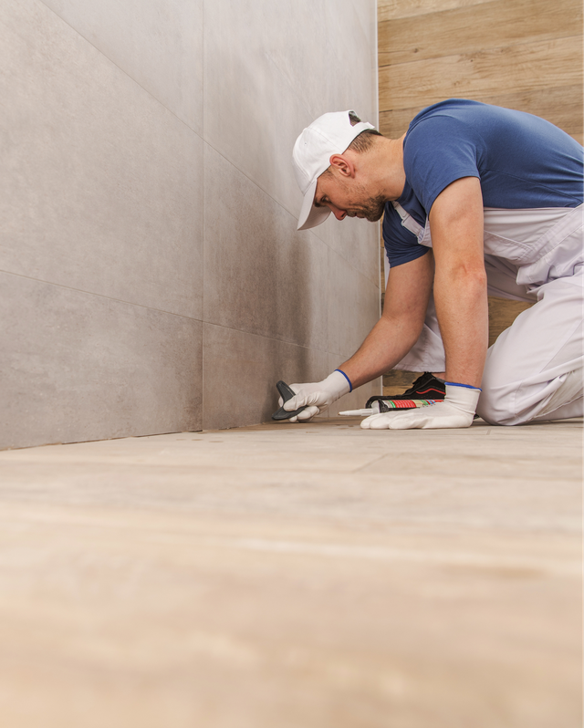 Worker installing flooring at base of a wall, kneeling with tools in a room