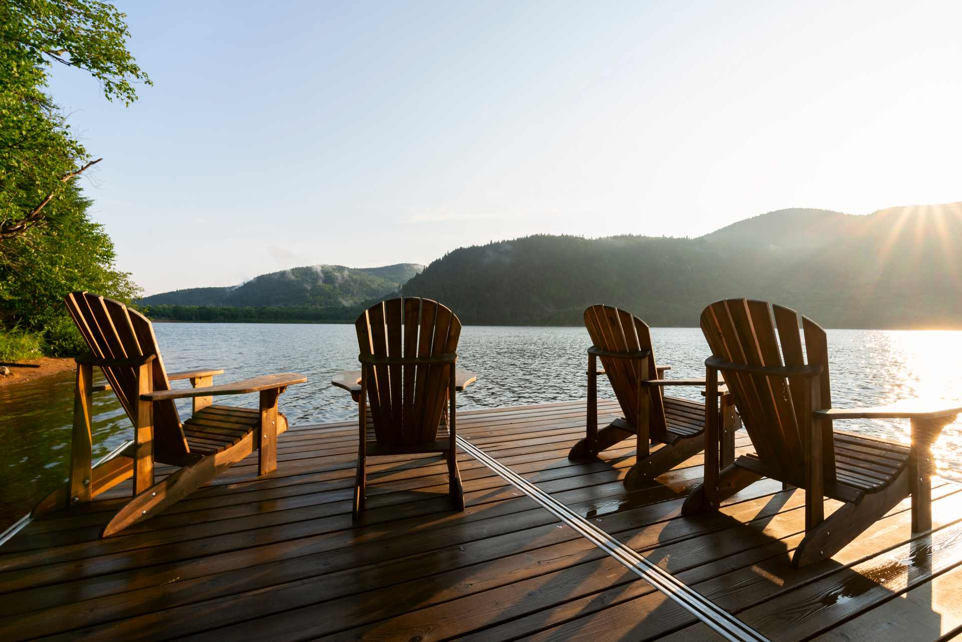 Wooden chairs on a lakeside deck at sunrise, with mountains and bright sun reflecting on the water.