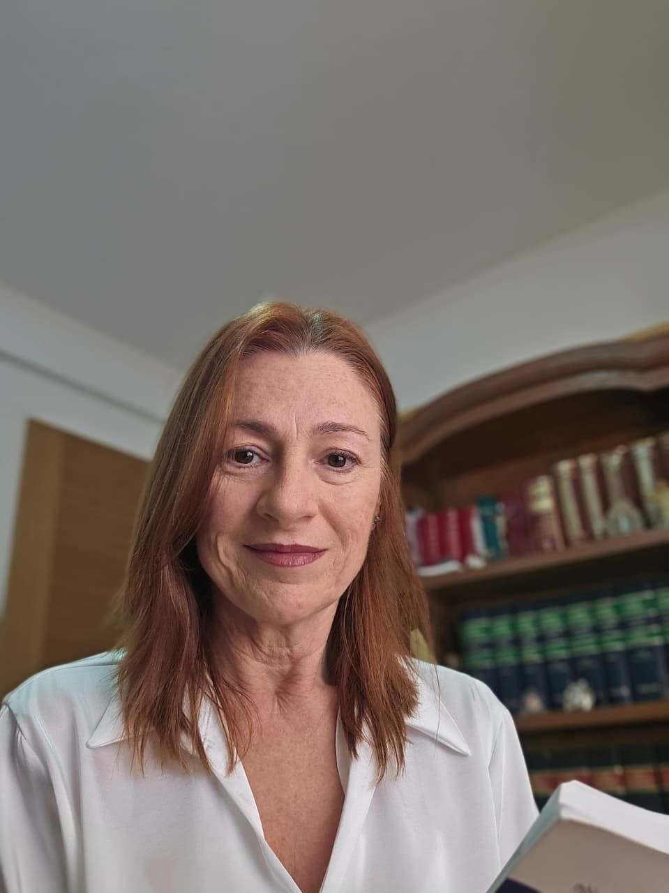 Mujer con blusa blanca de pie en el interior de una casa, cerca de una estantería, sonriendo a la cámara.