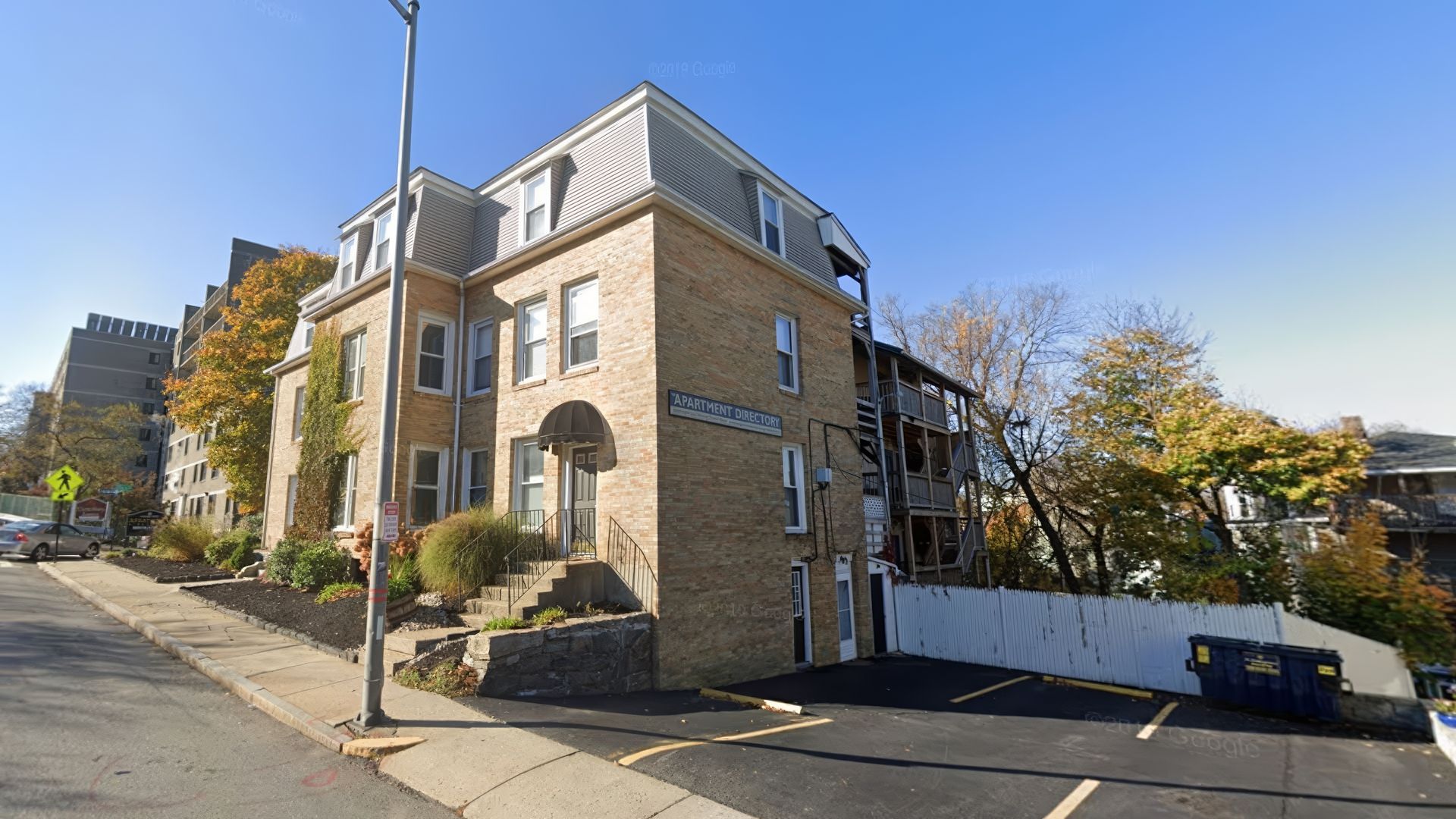 Brick apartment building on a street with parking. Trees and a blue sky are visible.
