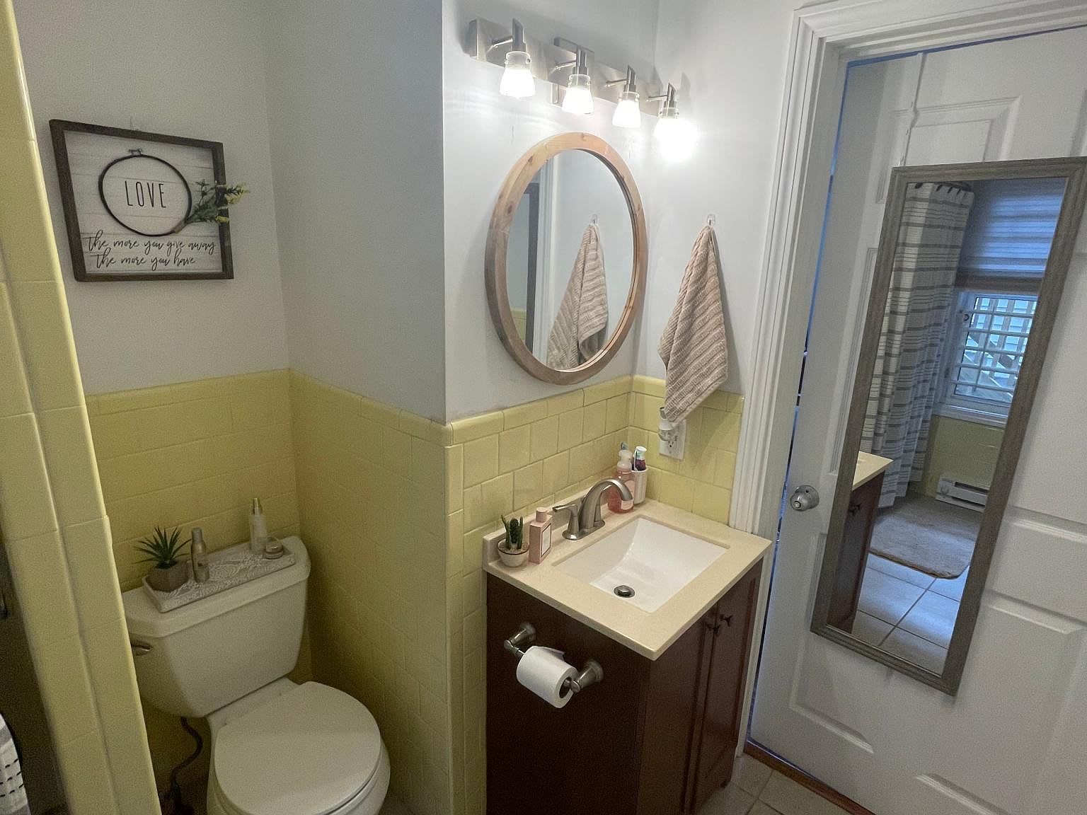 Small bathroom with yellow and white walls, toilet, vanity with round mirror, and a full-length mirror on the door.