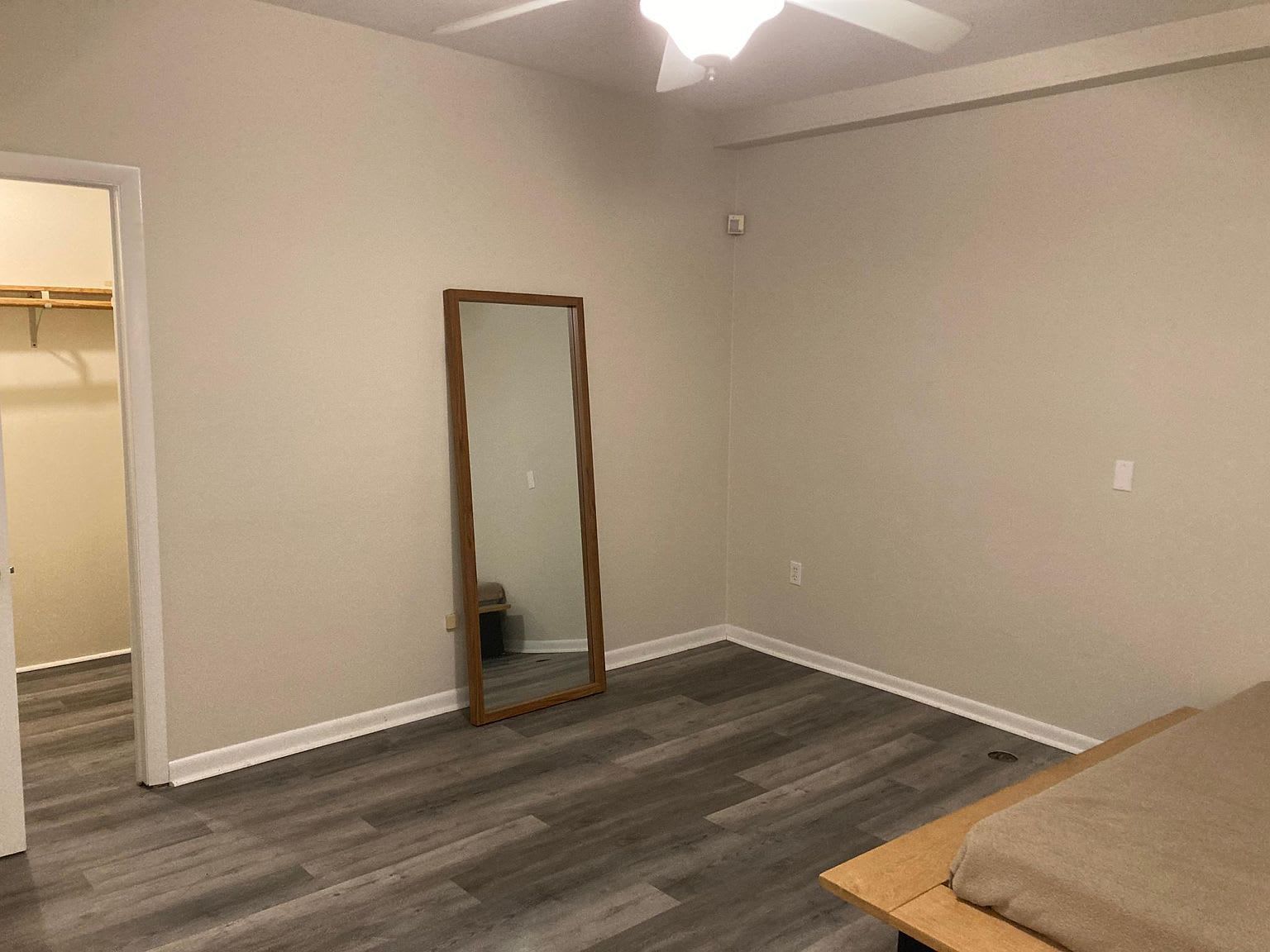 Empty bedroom with a full-length mirror, closet, and wood-look flooring. Walls are neutral, and a bed is in the corner.