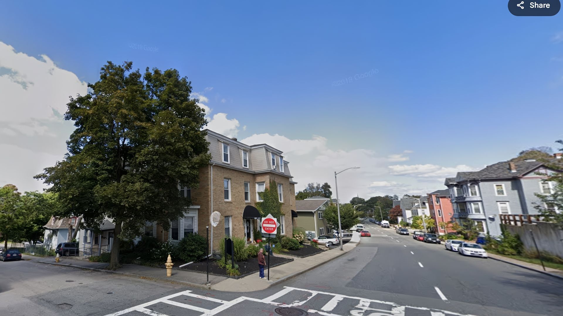 Street view of a two-story building with a distinctive roof, a tree, and a street with parked cars.