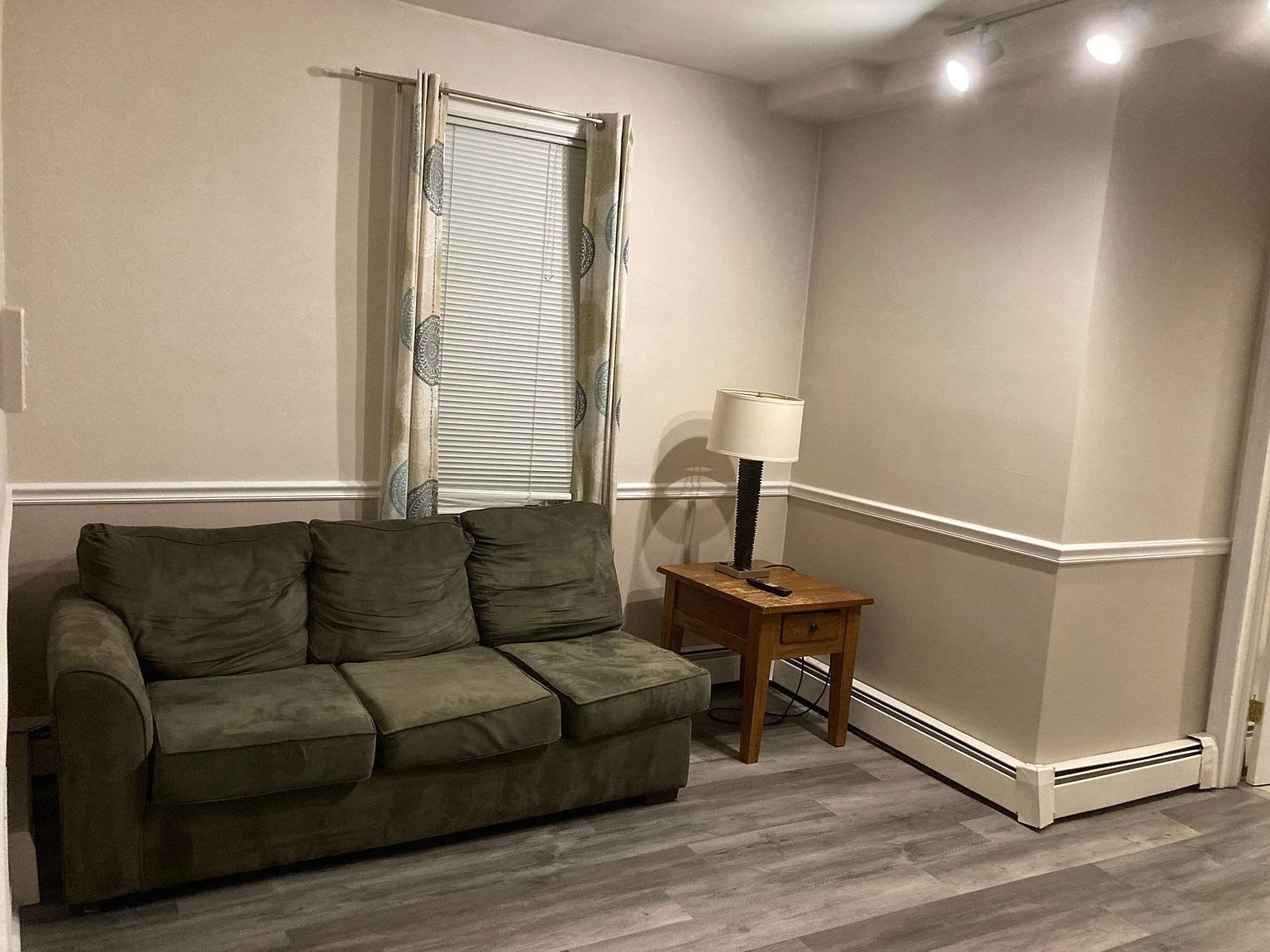 A living room with a green couch, wooden side table, and a window with blinds and curtains.