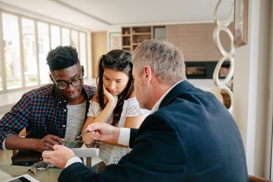 A financial advisor points to a document while consulting with a young couple at a table.