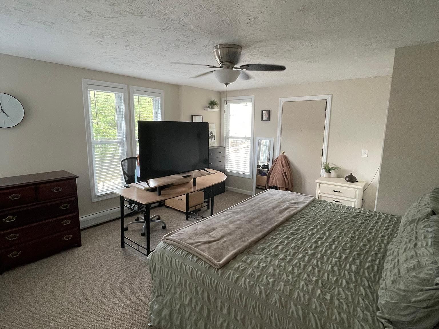 Bedroom with bed, desk with TV, dresser, windows, neutral colors.