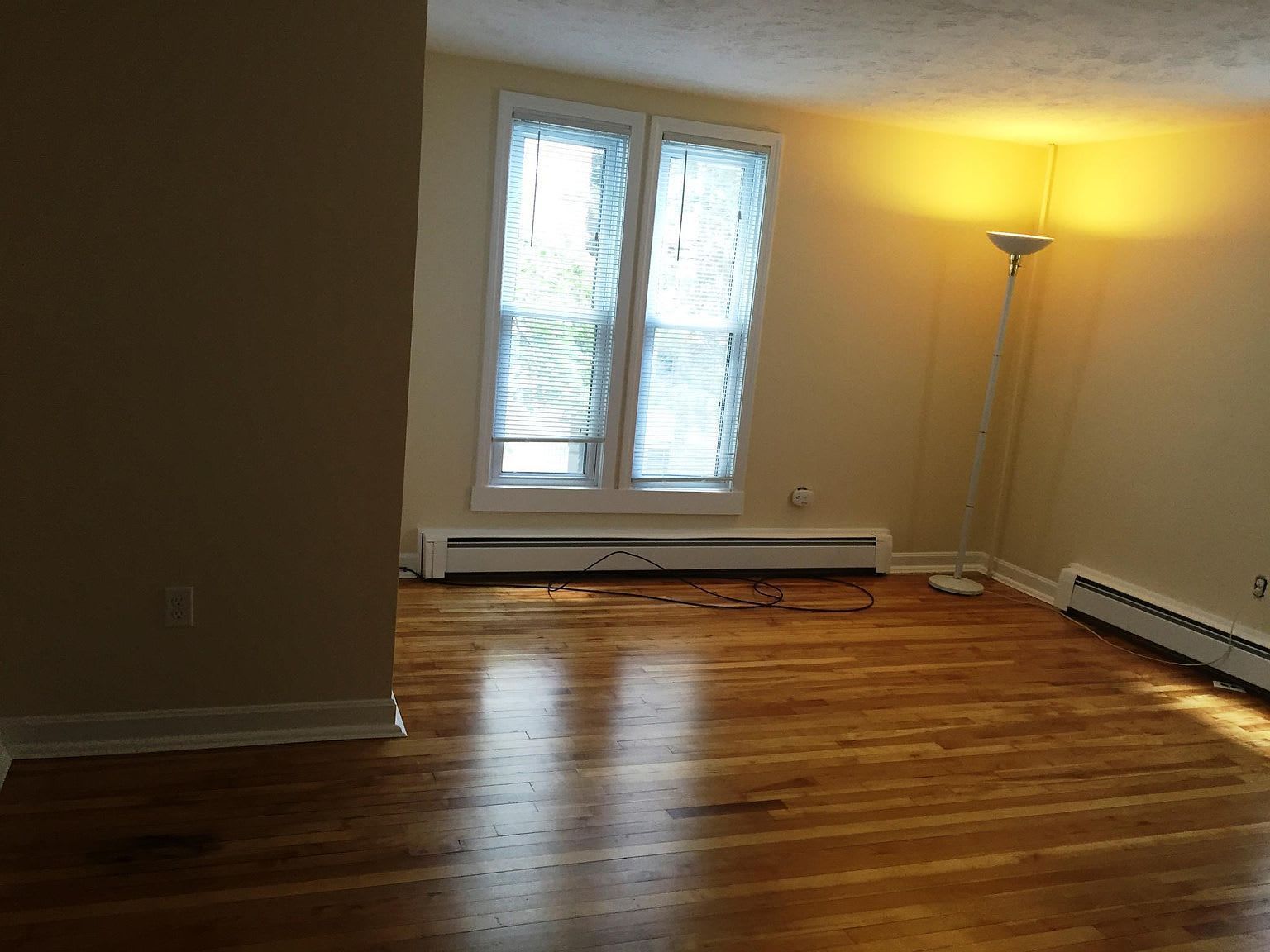 Empty room with hardwood floors, a window with blinds, and a tall lamp.