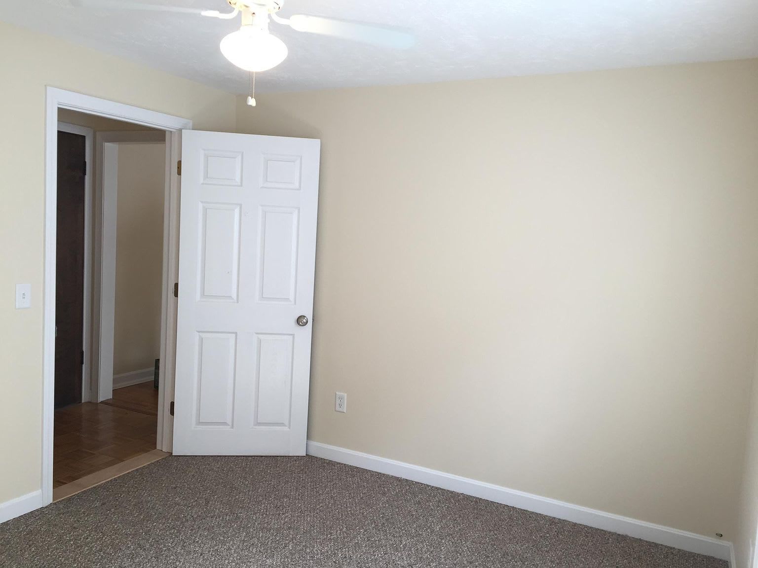 Empty bedroom with beige walls, white door, and carpeted floor. Doorway leads to another room.