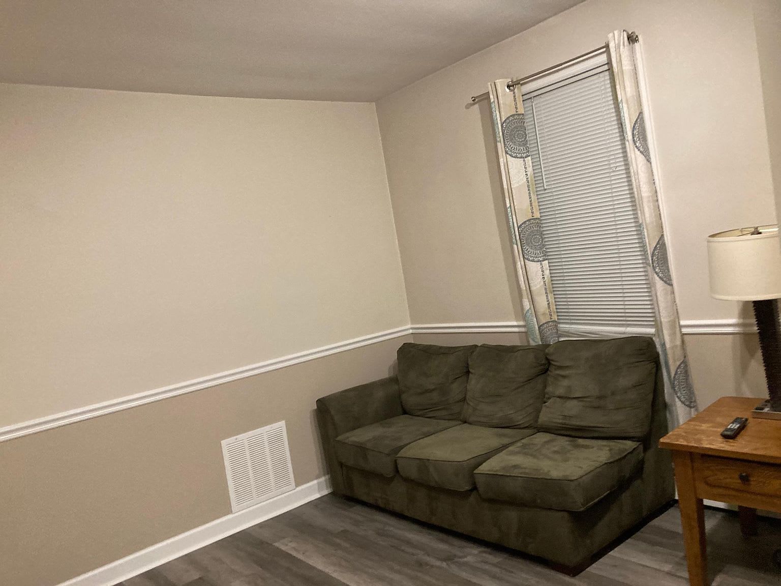 A small room with a green couch, window with patterned curtains, and a side table with a lamp.