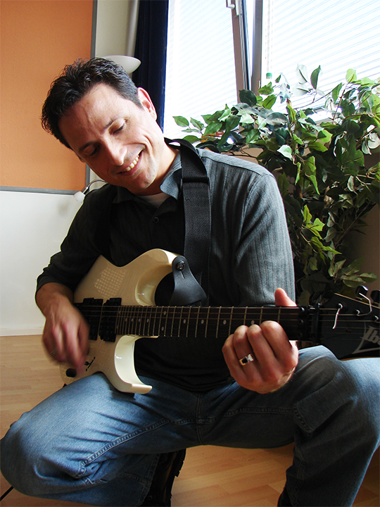 Al playing a white electric guitar, smiling, seated indoors.