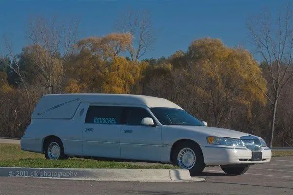 White hearse parked on a gray curb with trees and a blue sign visible.