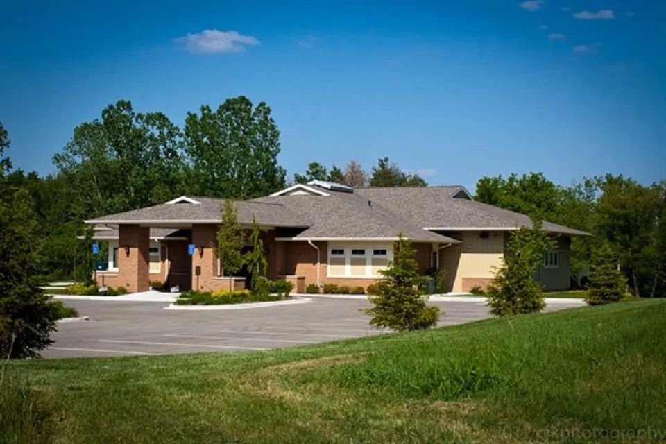 A one-story building with a gray roof and tan and brick facade, situated on a green lawn with a parking lot.