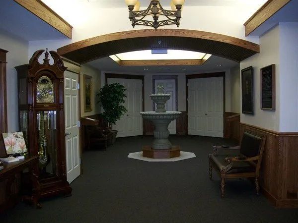 Interior foyer with fountain, grandfather clock, doors, and chandelier. Dark carpet, wood trim, and lighting.