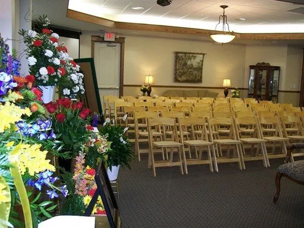 Funeral home interior with rows of chairs, floral arrangements, and a framed picture.