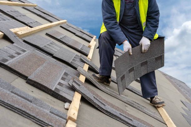 Roofing contractor in safety gear installs asphalt shingles on roof, with blue sky background.