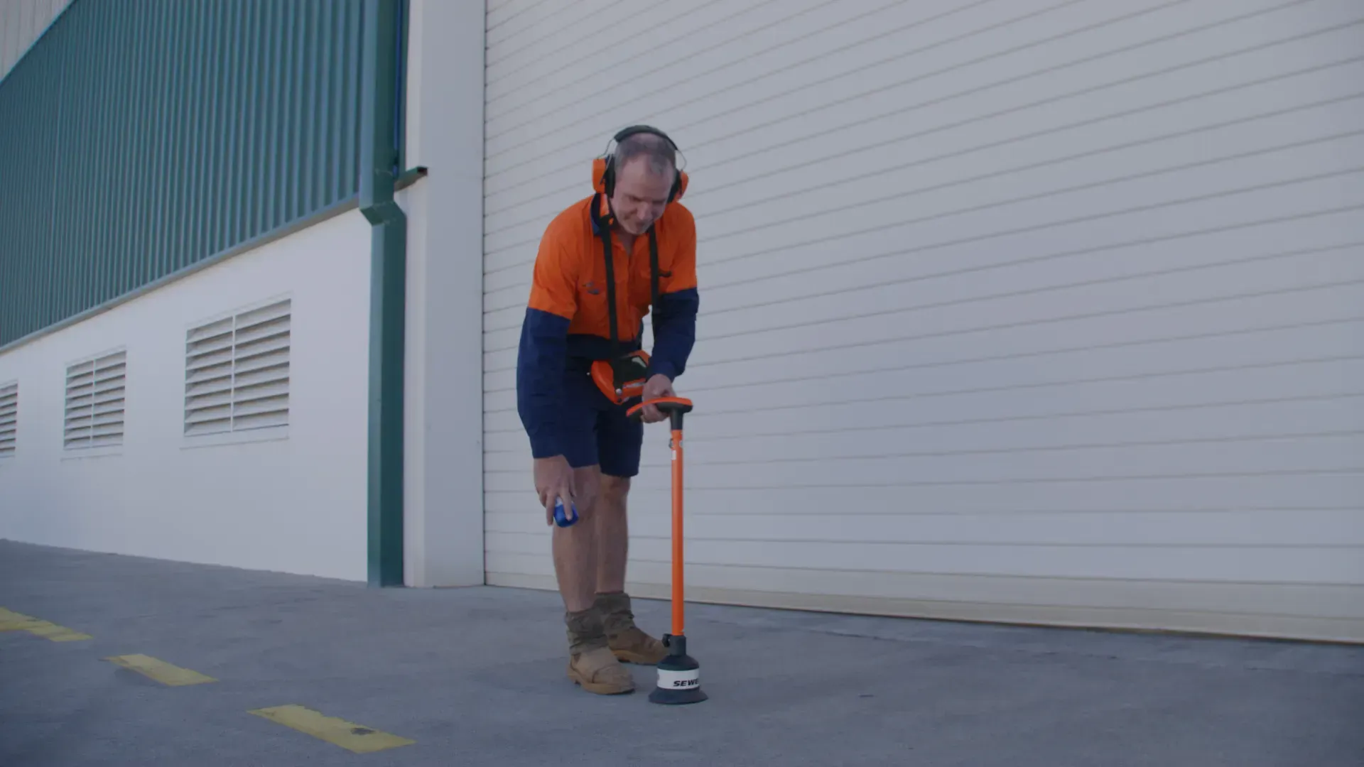 Man using detection tool outdoors, wearing safety gear, near a building.