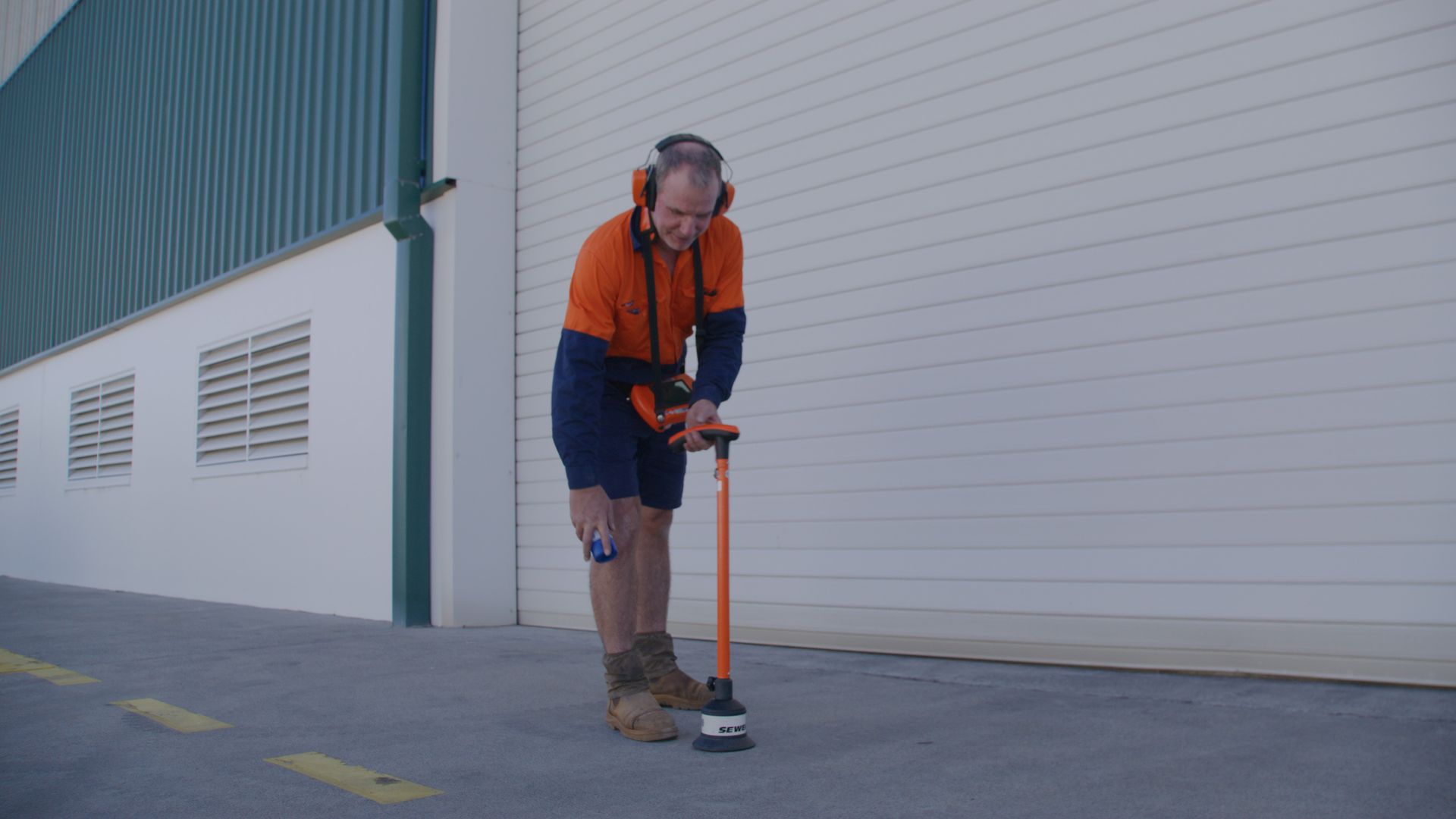 Worker in orange shirt uses a tool on pavement near a building.