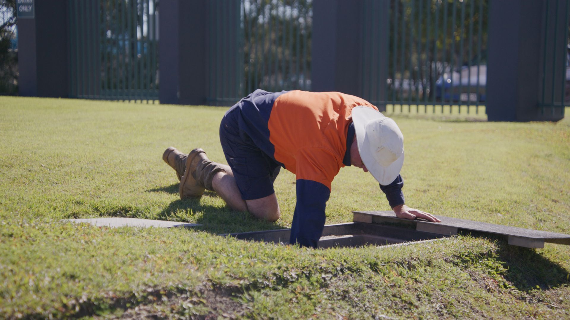 Person in work clothes kneels, removing a cover from an access panel in a grassy area, sunny day. — Commercial Leak Detection in Gold Coast, QLD