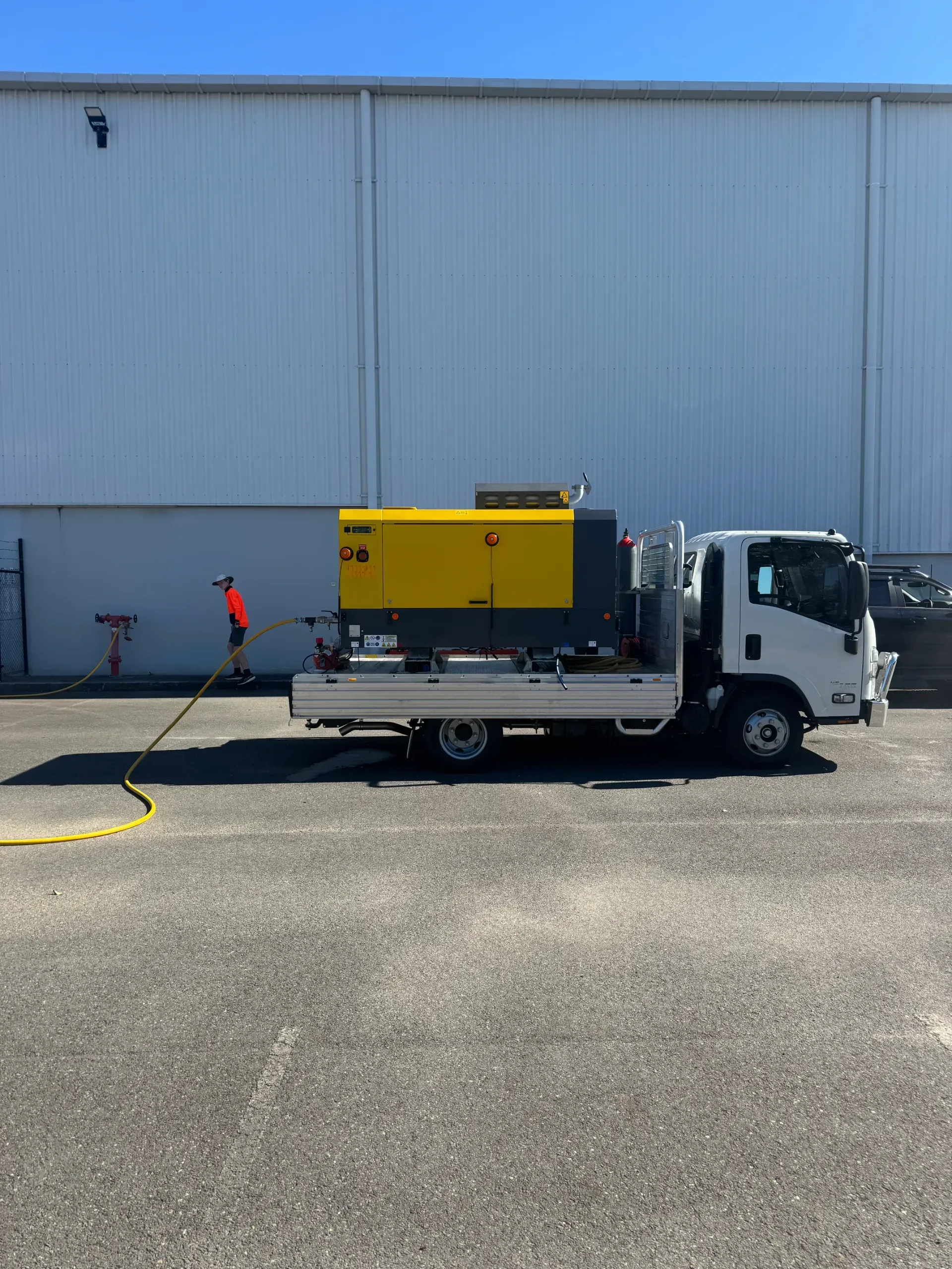 A truck with a yellow and gray machine on the back, person using a hose, building background.