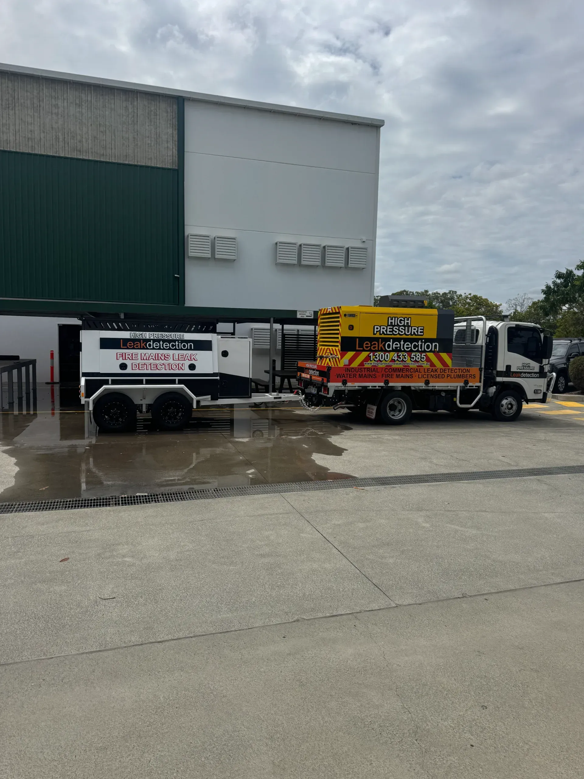 Two utility trucks parked near a building, water on the ground.