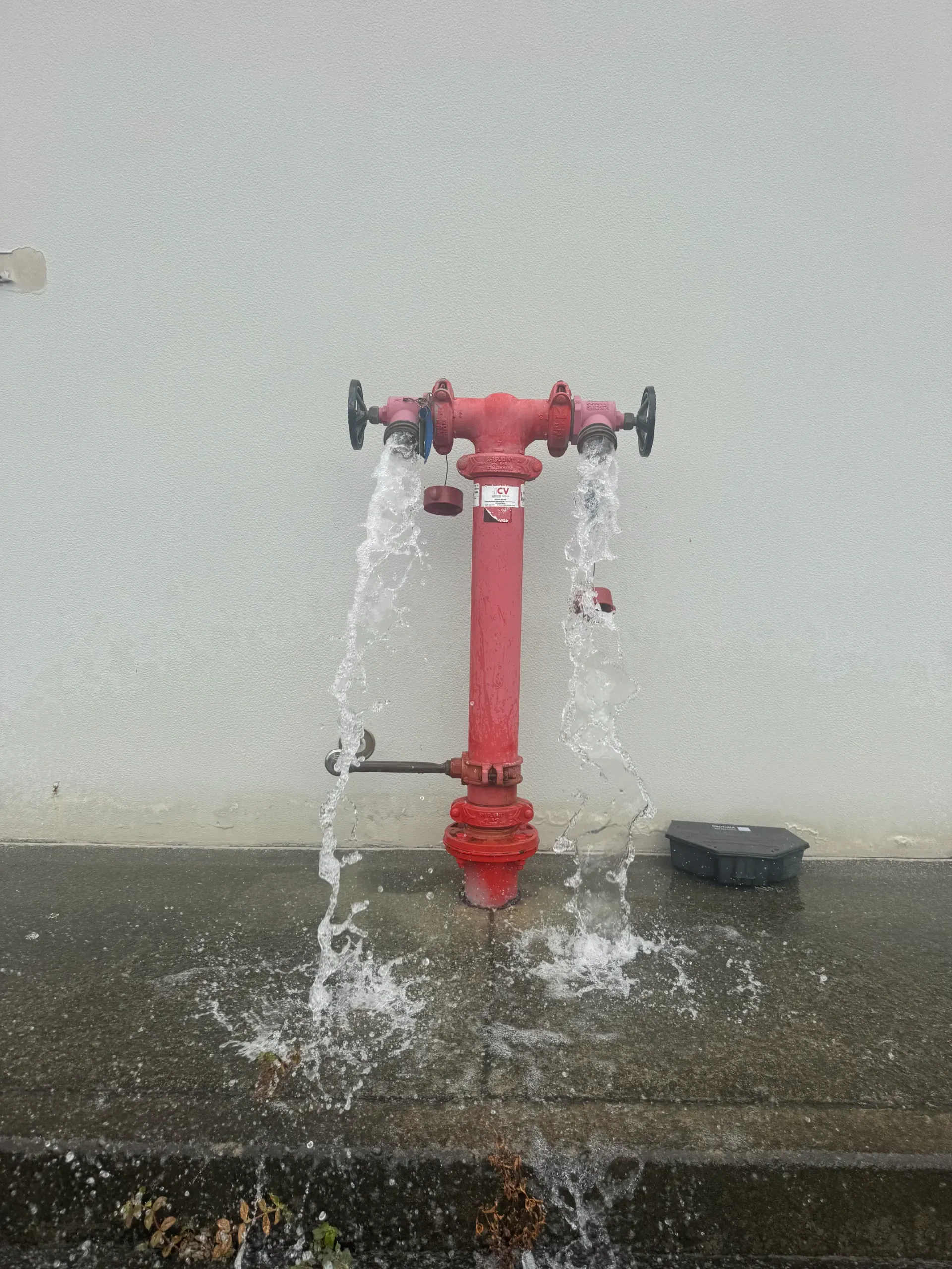 Red fire hydrant spraying water against a light-colored wall.