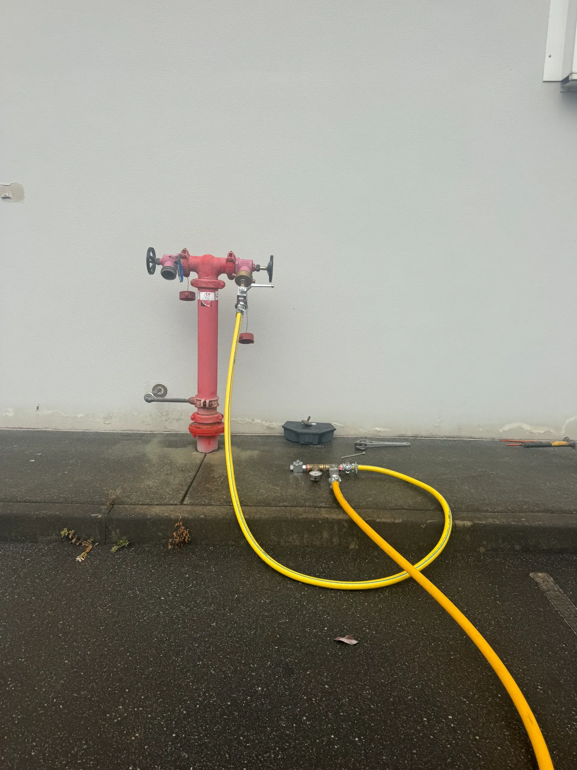 Red fire hydrant with a yellow hose connected, on wet pavement next to a white wall. — Commercial Leak Detection in Molendinar, QLD