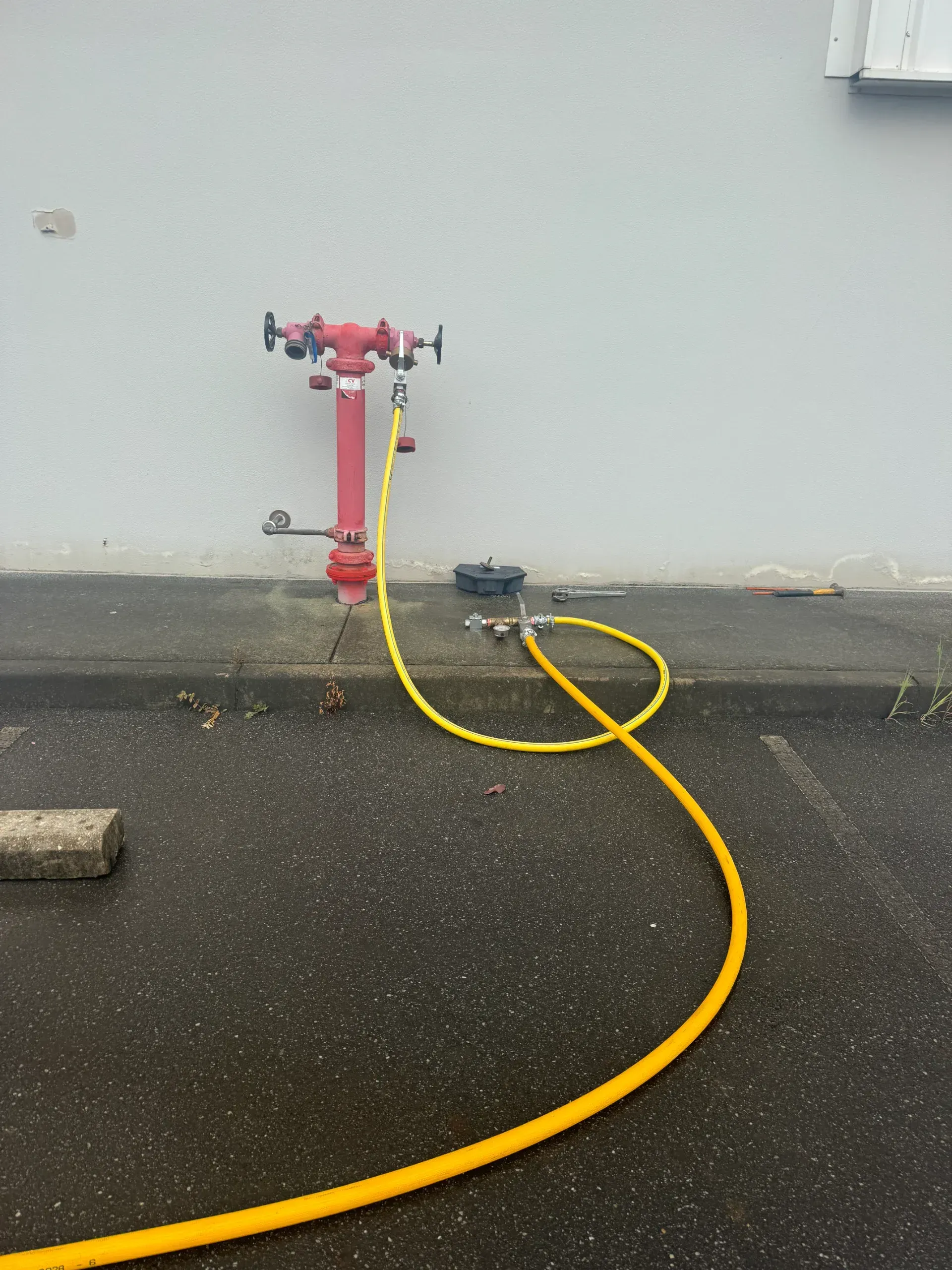 Red fire hydrant connected to a yellow hose on a wet, gray surface near a building.