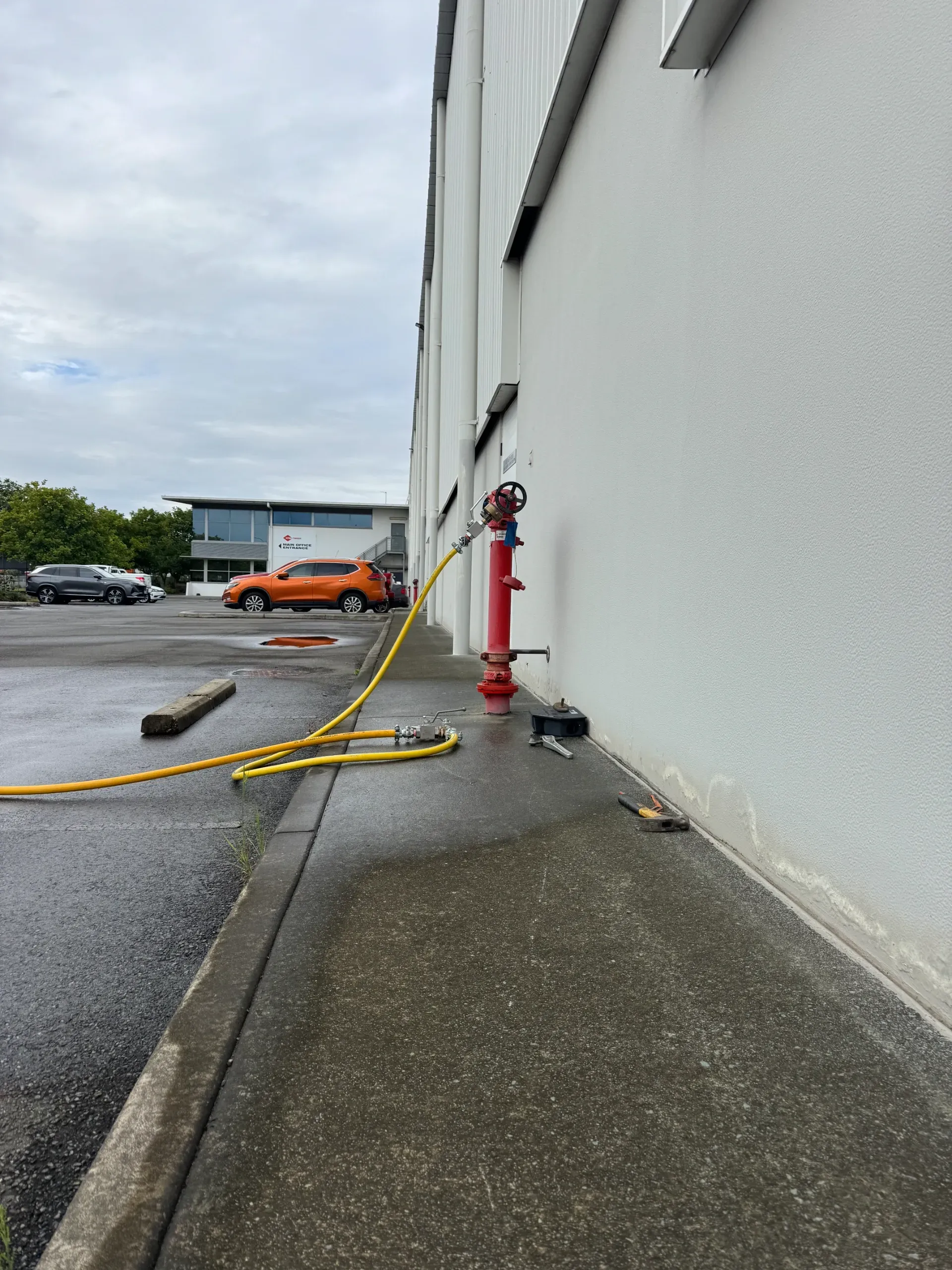 A red fire hydrant on a white building with a yellow hose laid out. Car in background. — Commercial Leak Detection in Ballina, NSW
