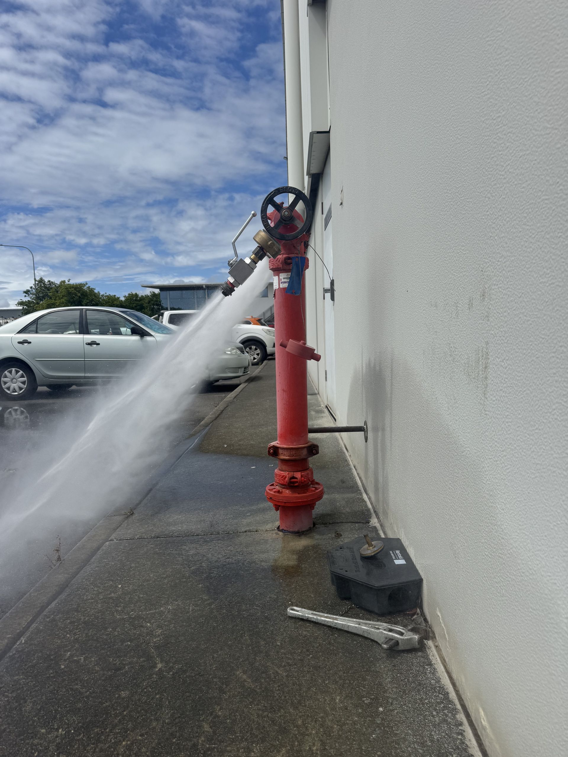 Red fire hydrant spewing water next to a white building. A car is parked nearby.