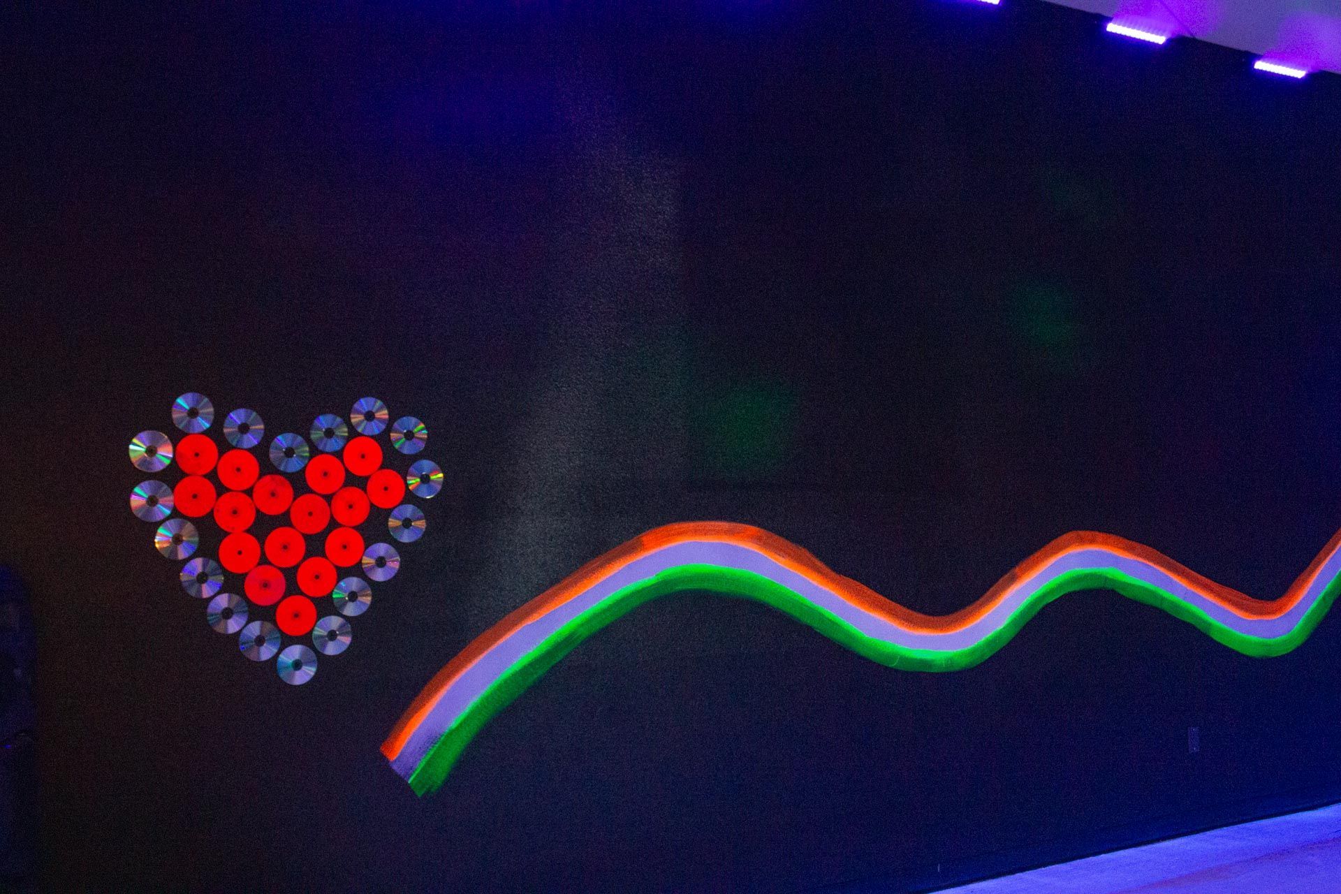 Heart made of red circles and silver discs, wave-like design with orange, green, and blue lines on a black wall.