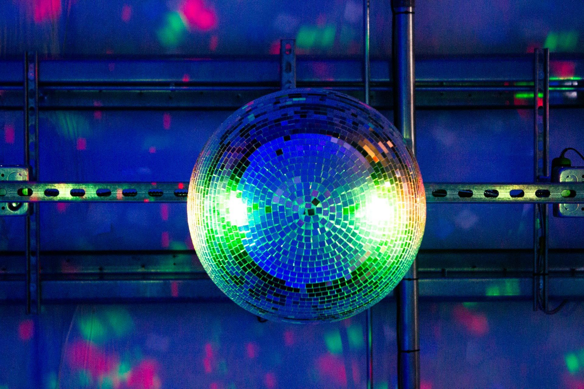 Glittering disco ball hangs under metal beams, reflecting blue, green, and pink light.