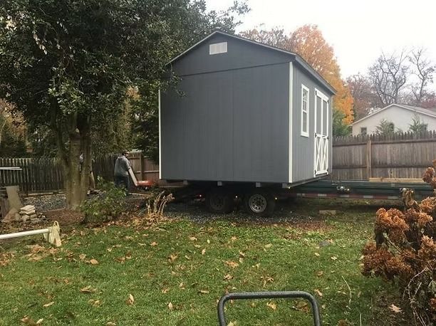 Gray tiny house on a trailer in a grassy backyard with trees and a wooden fence.