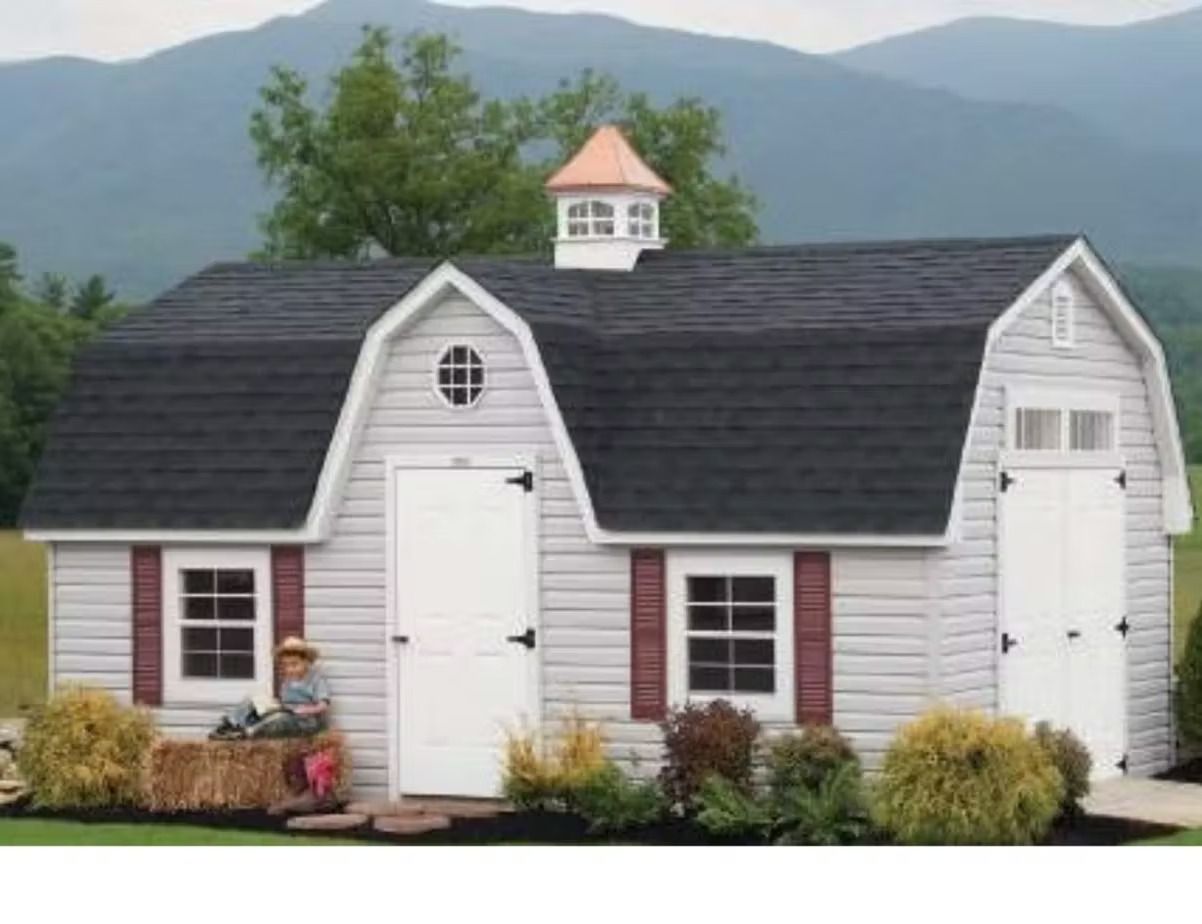 White shed-style house with black roof, red shutters, and flower beds in front of mountains