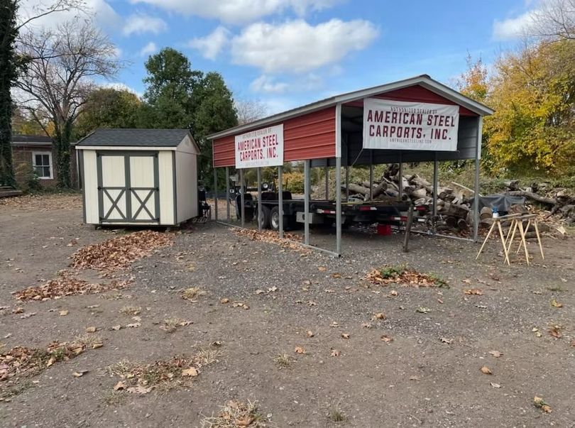 Outdoor farm stand with red canopy, white shed, and pumpkins on gravel under autumn trees