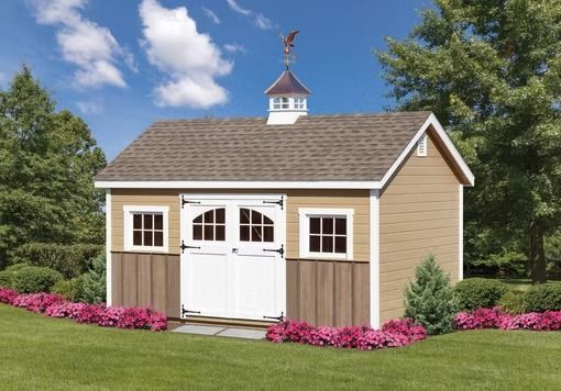 Small tan shed with white double doors and windows in a green yard, framed by pink flowers and trees.