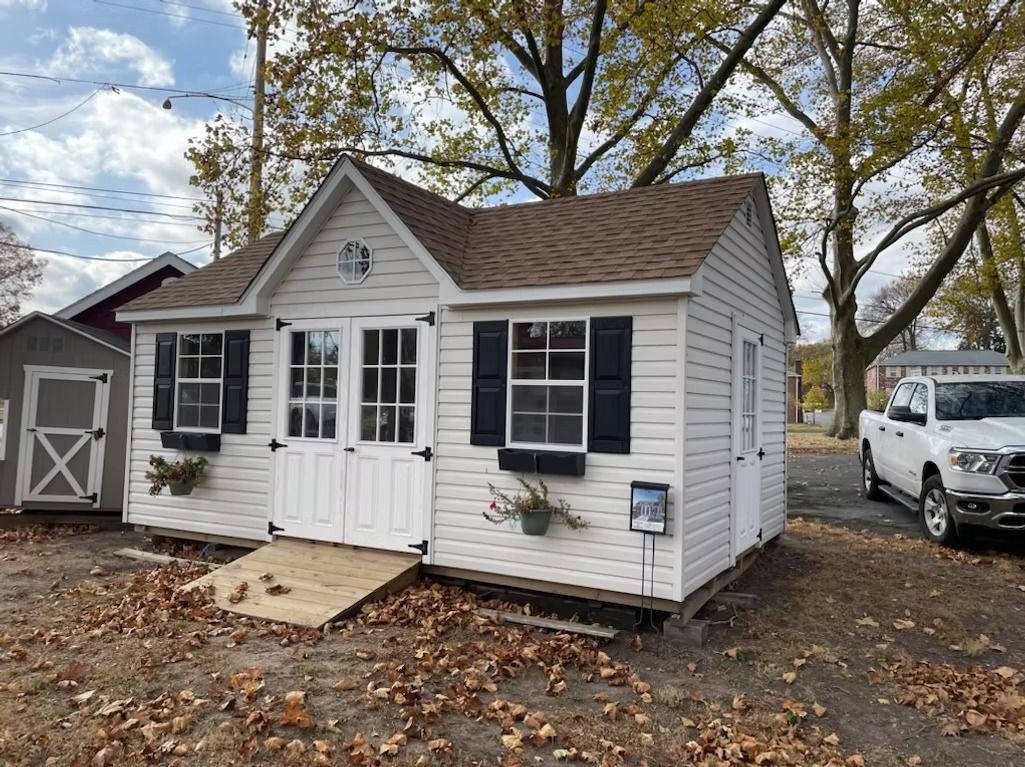 Small white cottage with black shutters and a front porch, set on a leaf-covered lot with trees and parked vehicles.