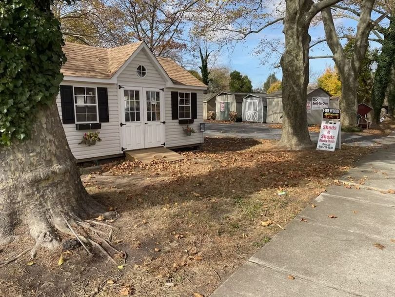 Small white house with black shutters beside a sidewalk, surrounded by trees and a yard in autumn.