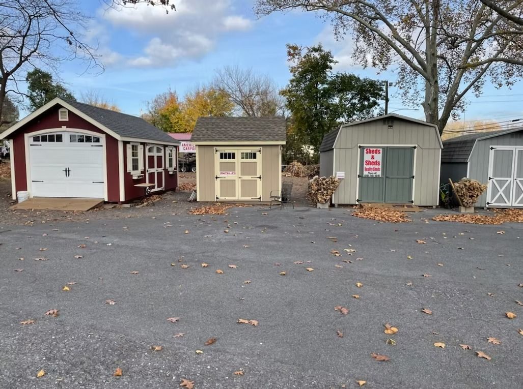 Small row of sheds and a garage on a paved lot, with autumn trees in the background.
