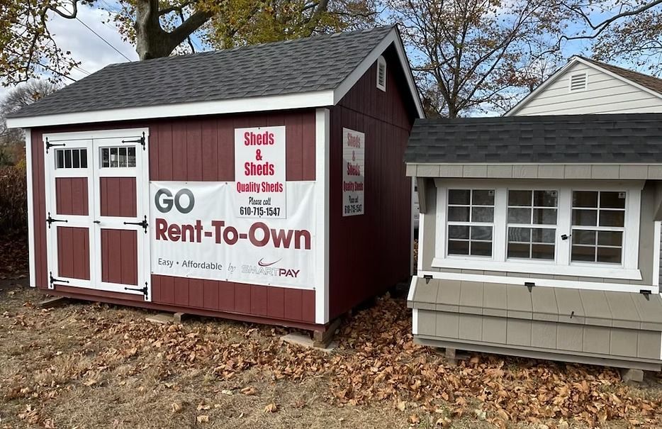 Small maroon and white rent-to-own shed beside a gray house, with “GO Rent-To-Own” sign.