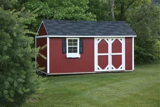 Red garden shed with white trim and double doors on a grassy lawn beside trees.